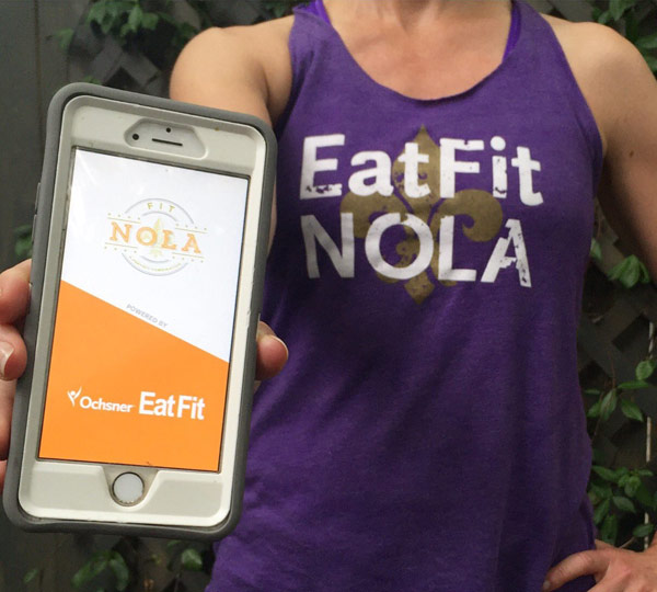 Lady with Eat Fit NOLA shirt holding smartphone with Oschner Eat Fit Logo on Screen